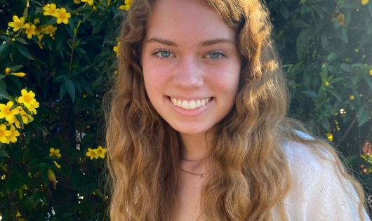 A blonde young woman with curly hair smiles at the camera.