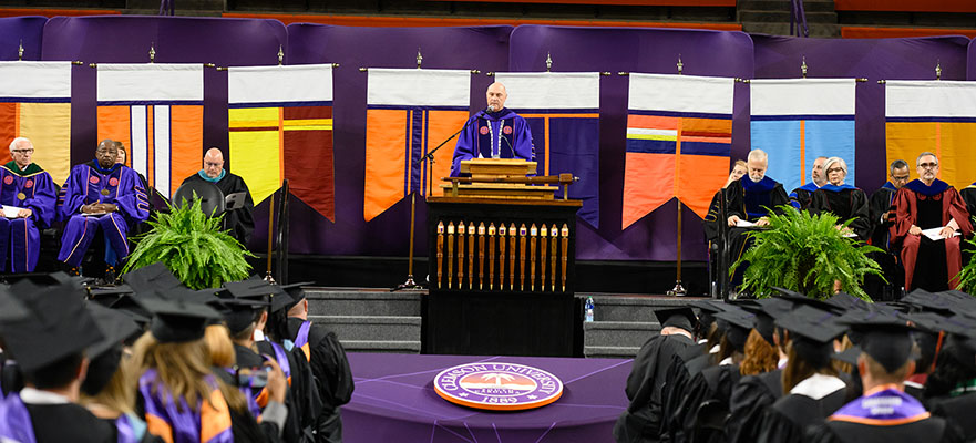 President Clements stands on stage at graduation with the College Banners displayed behind him. 