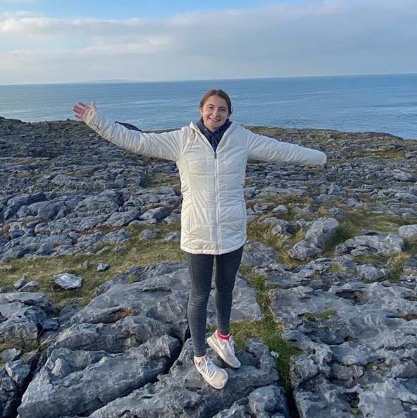 Megan Noonan smiling and standing with her arms outstretched on a rocky beach. 