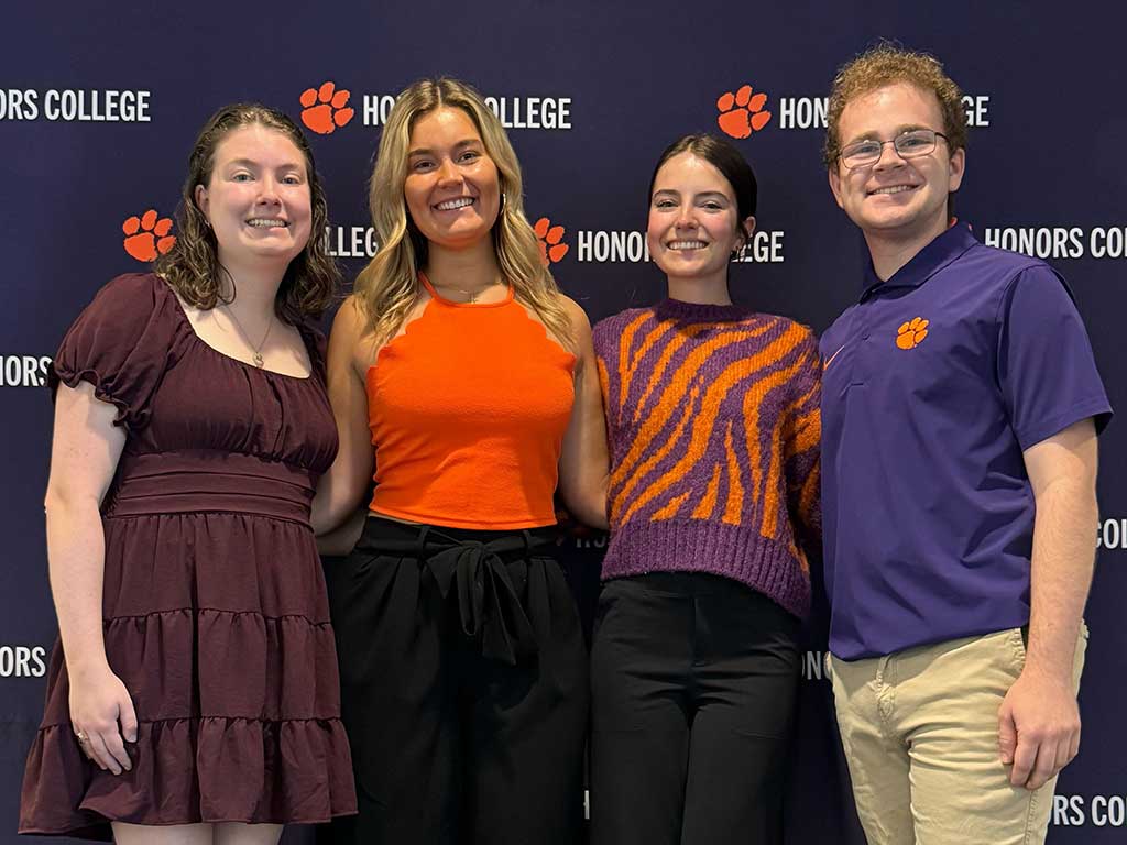 Four students standing and smiling in front of an Honors College backdrop.