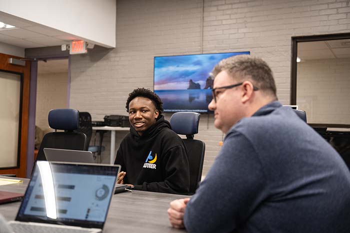 A student sitting at a table talking to a faculty member, with an open laptop beside them. 