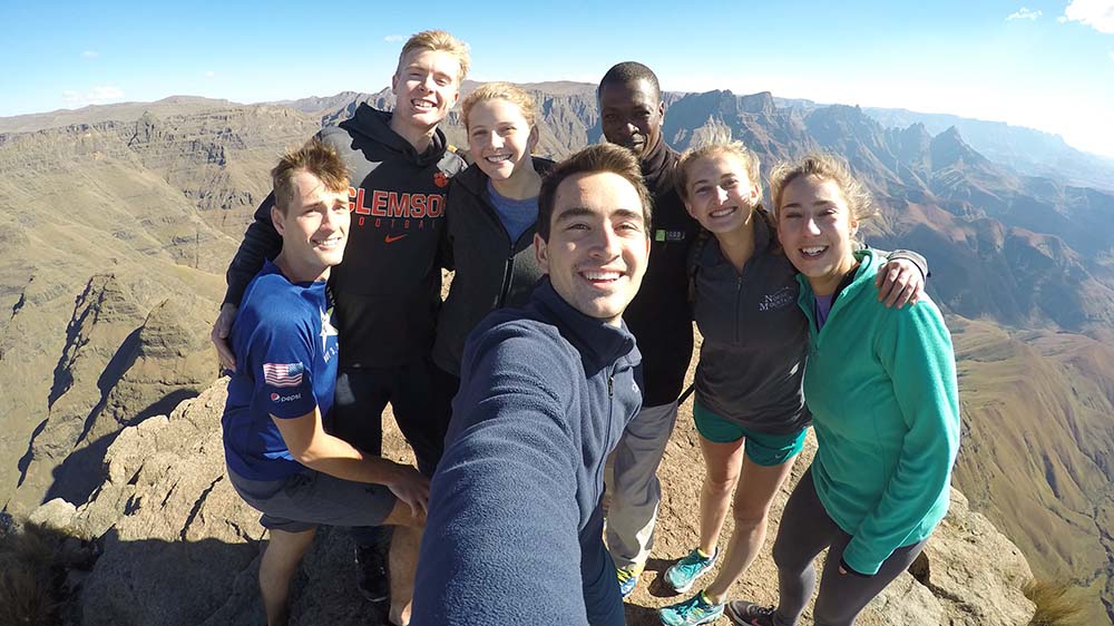 A student selfies with six peers at the top of a large mountain range