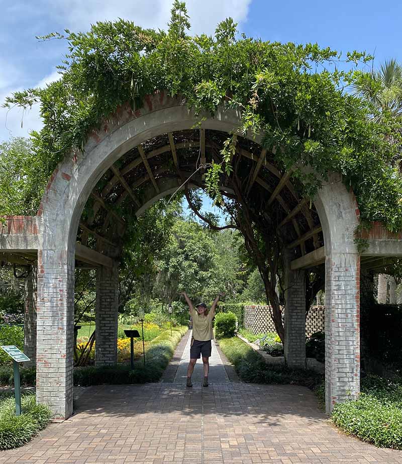 Student standing beneath stone arch covered with vines at Moor Farms Botanical Gardens.