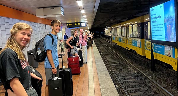 Students standing in a line with their suitcases by their feet while waiting for a subway train