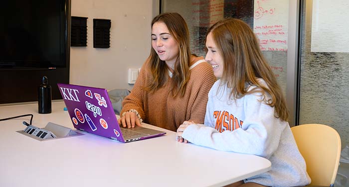 Two Honors students smiling and looking at a laptop in an Honors study room.