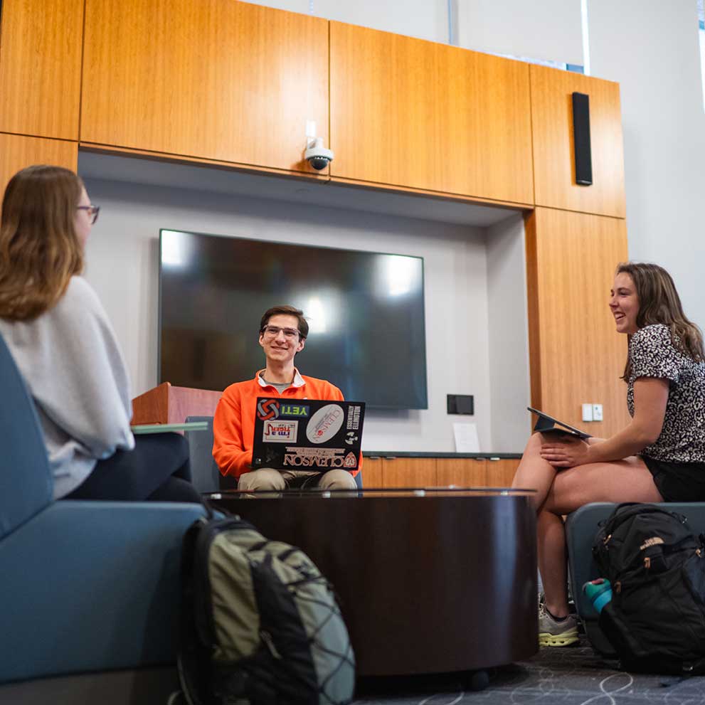A male Honors student sitting in the Great Hall talking to two other students, who are also seated.