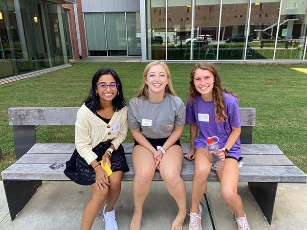Three female students sit on a bench and smile outside the Honors Center