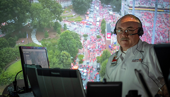Fire Chief in front of the Command Center wide screen showing live footage of the crowds at a football game.