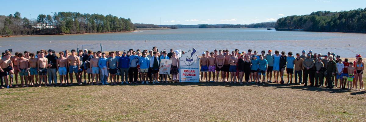 Large group of participants lined up on a lakeshore for a Polar Plunge event, with a banner and mascot sign in the center.