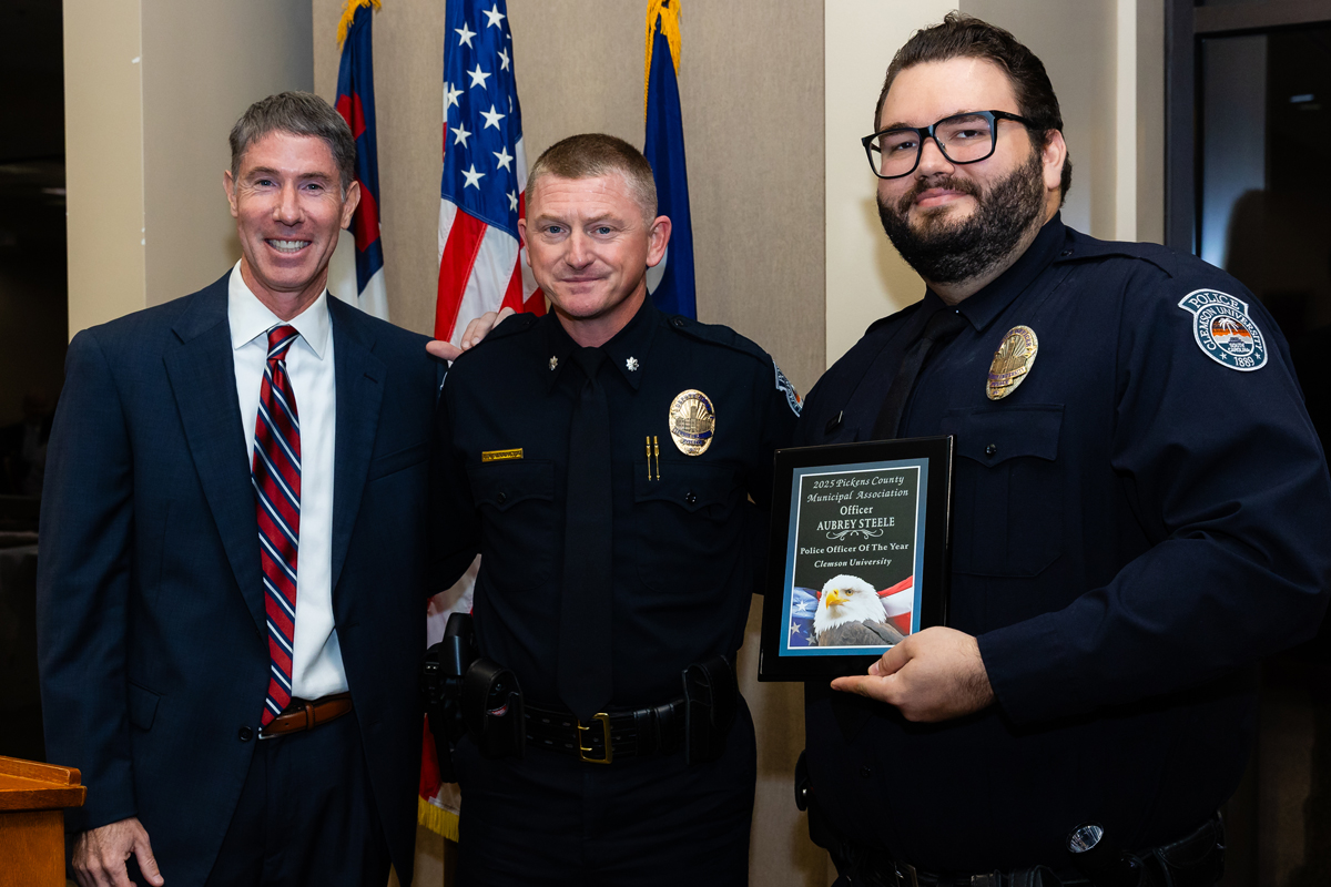 Three men pose for a photo at an awards event, with a police officer holding a plaque recognizing a Police Officer of the Year.