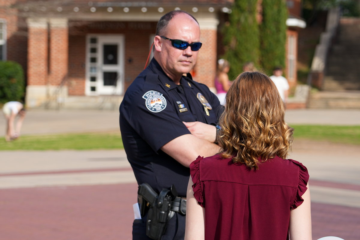 Police officer in uniform talking with a woman outdoors on a campus walkway.