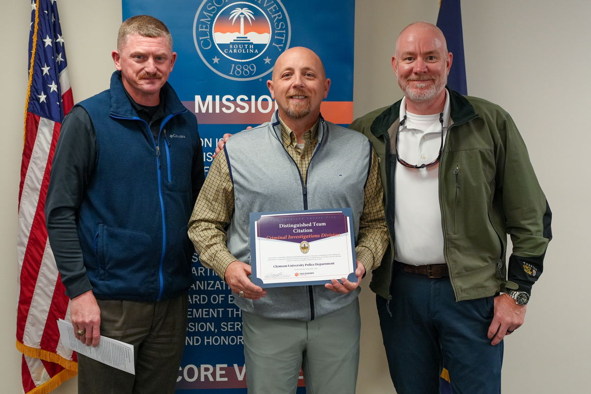 Three men pose with a Distinguished Team Citation certificate in front of a Clemson University Police Department banner and U.S. flag.