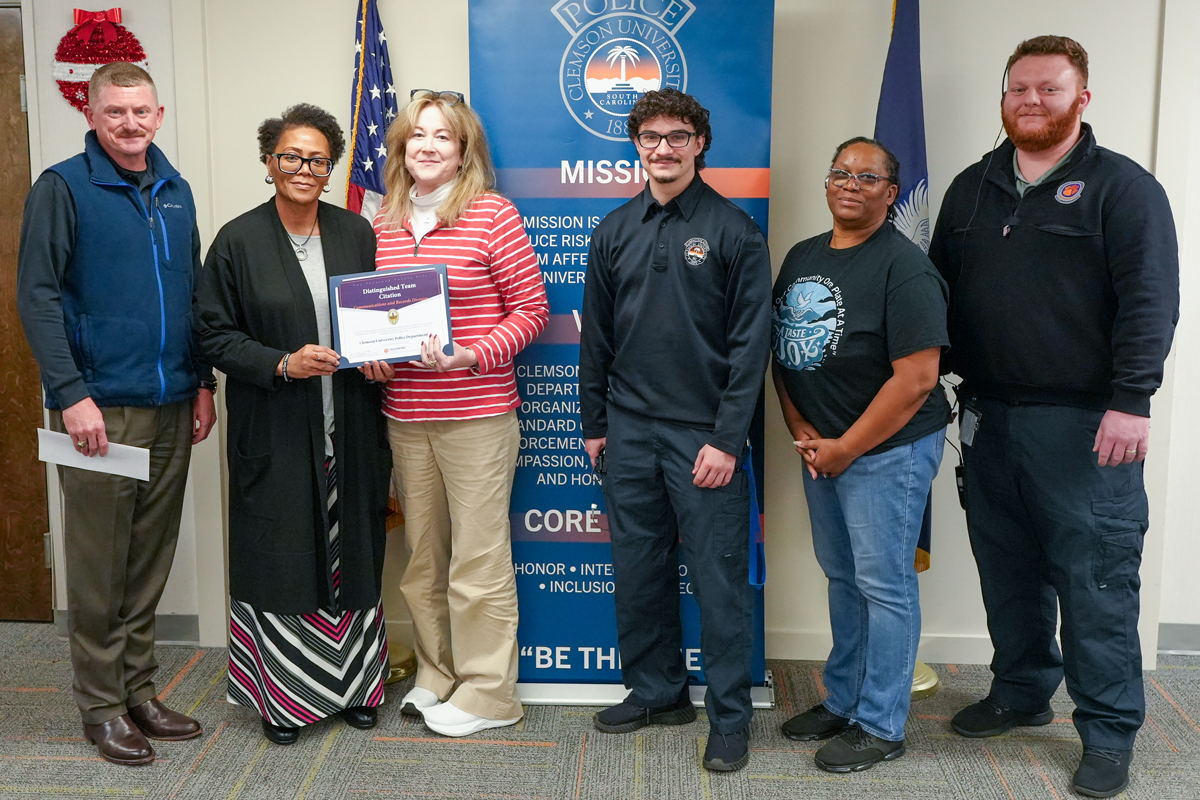 Group of six people posing indoors, with two holding an award certificate in front of a Clemson University Police banner and flags.