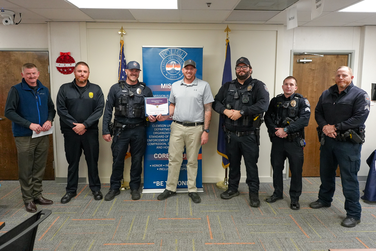 Group of police officers and staff posing indoors, with one person holding an award certificate in front of a department banner.