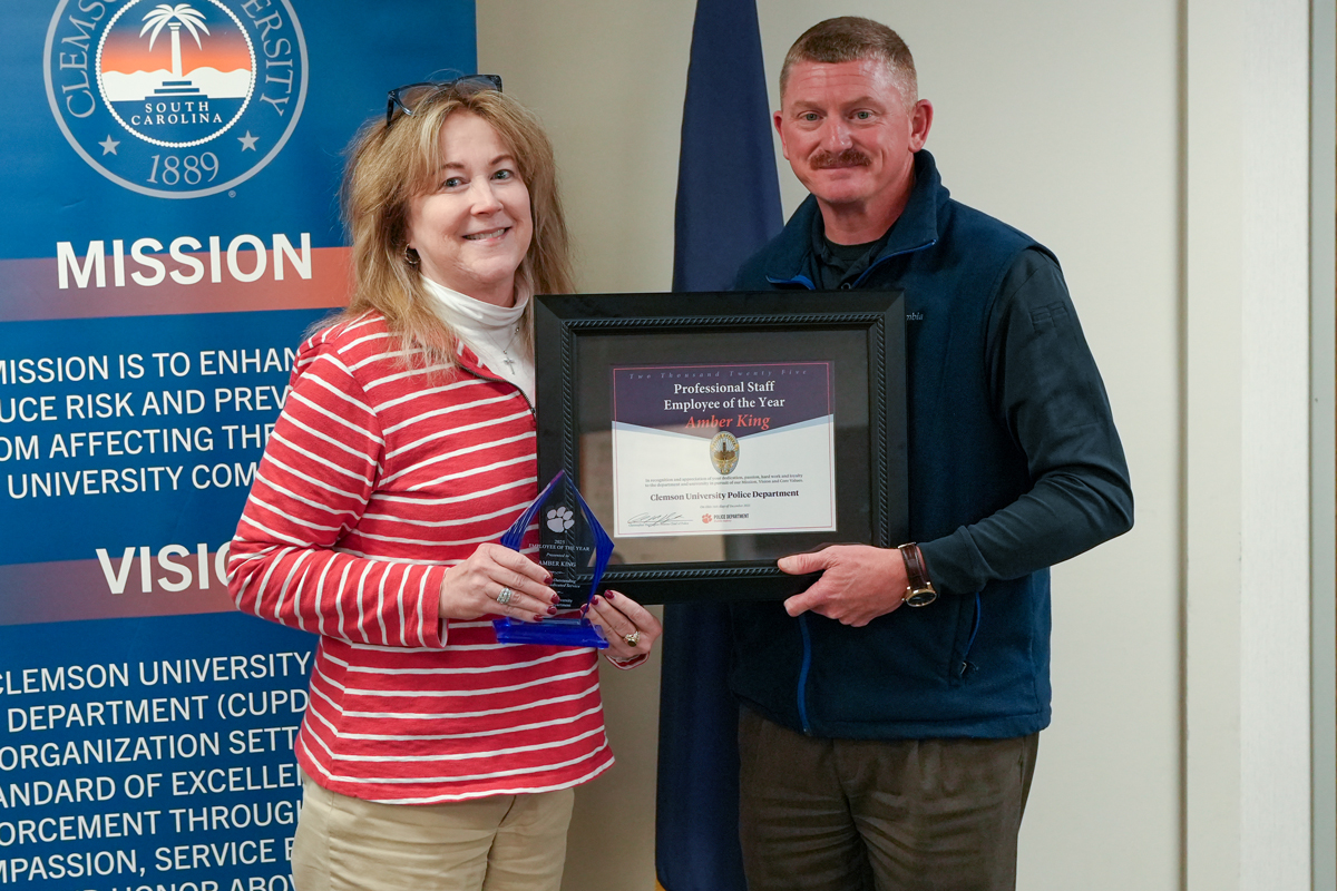 Two people pose holding a framed “Professional Staff Employee of the Year” certificate and a glass award in front of a Clemson University Police Department mission banner.