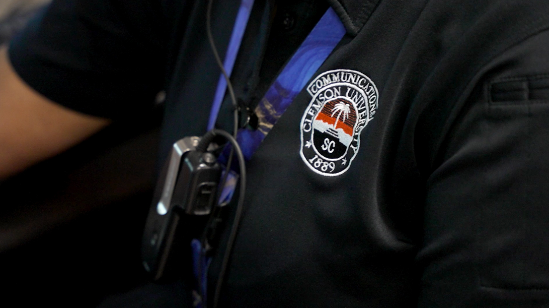 Closeup of a Clemson University communications center logo and a radio on the black shirt of an employee.