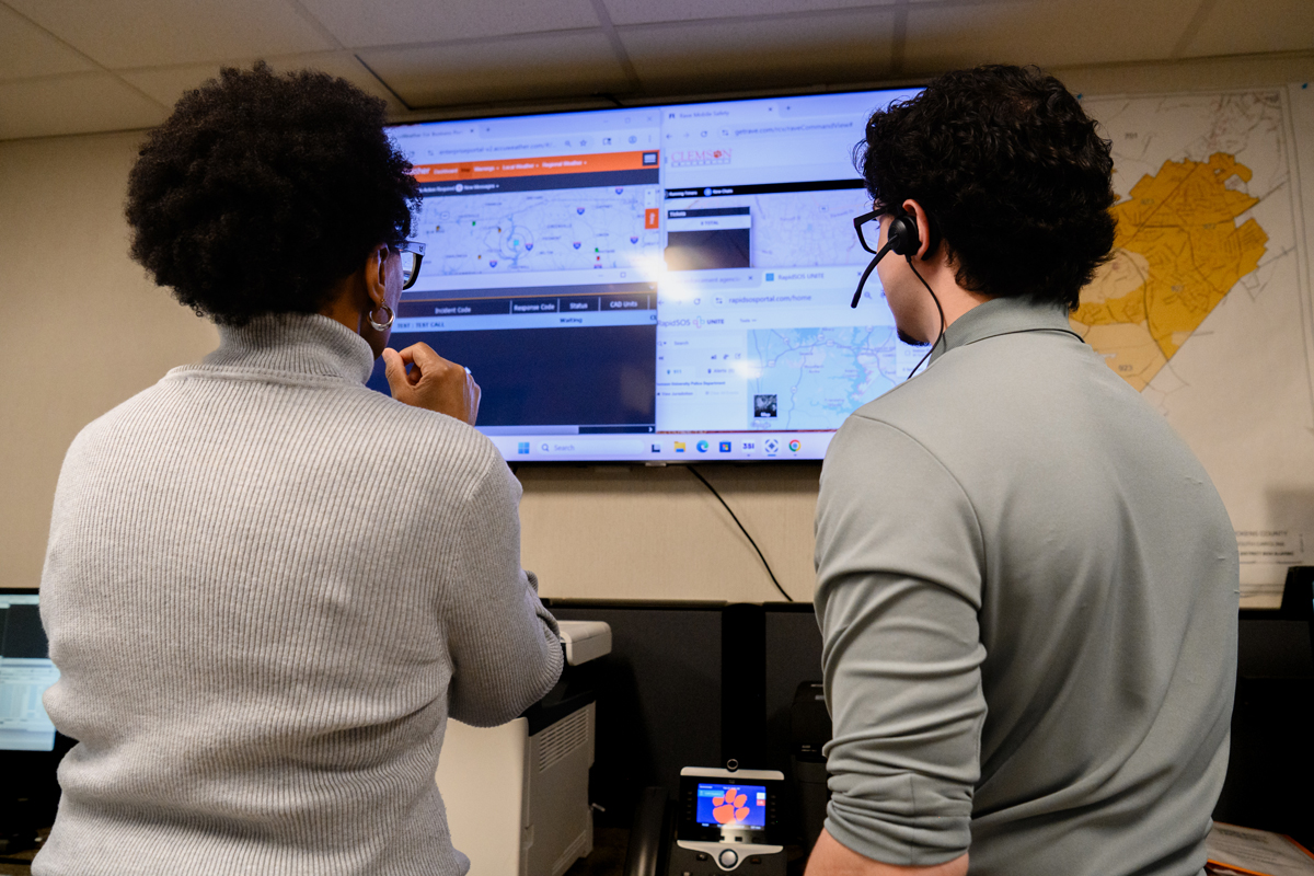 Two colleagues view a large wall-mounted monitor displaying maps and dispatch software in an operations center.