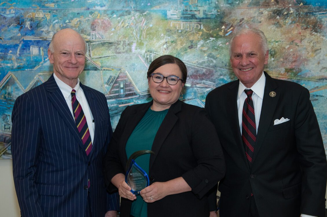 Three people posing together indoors, with the woman in the center holding a glass award in front of a colorful mural.