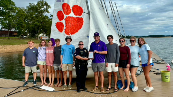 Group of teens and two adults standing on a dock by a sailboat with a large orange paw print on the sail, holding an award plaque.