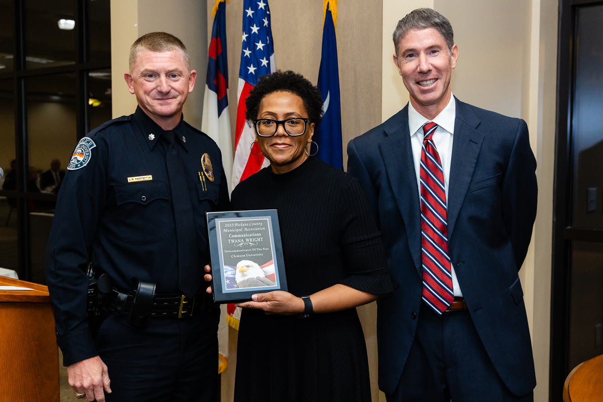 Woman holding an award plaque poses with a police officer and a man in a suit in front of U.S. flags.