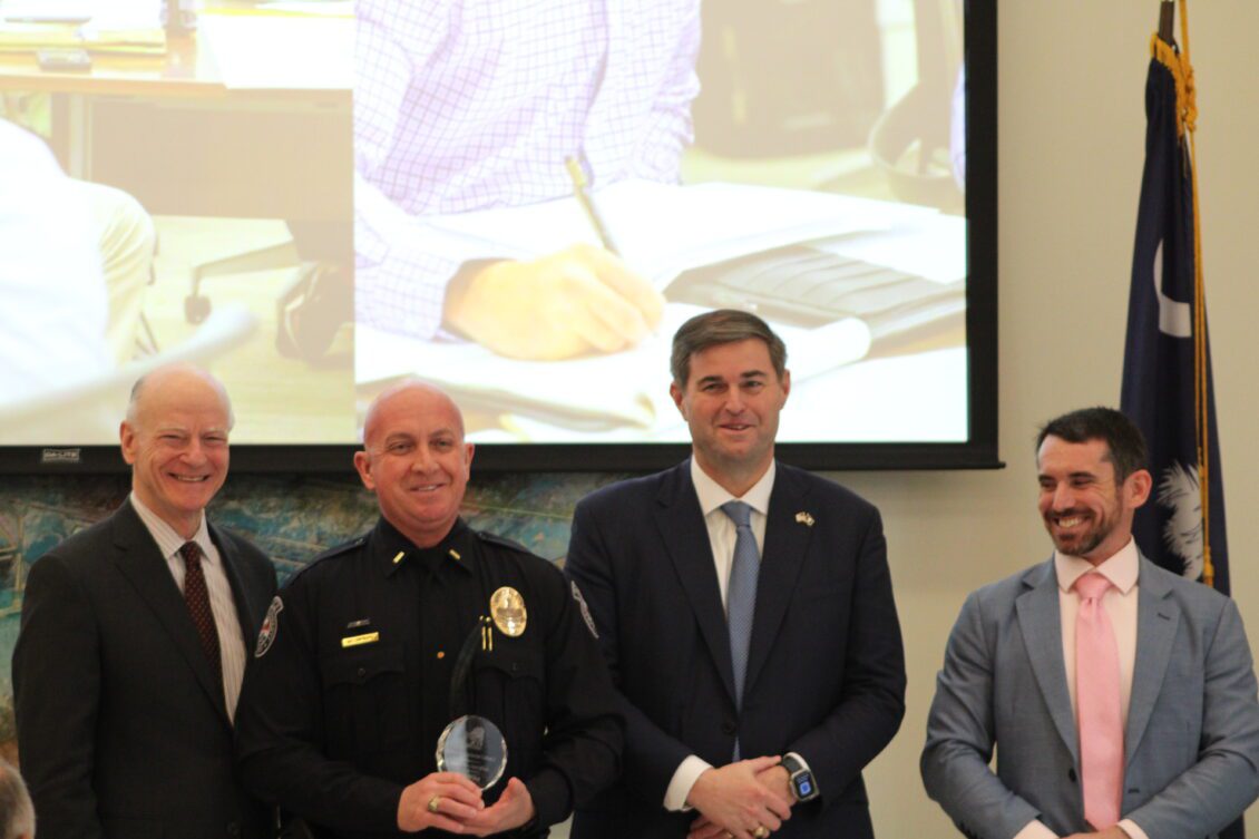 Four men at an indoor ceremony, including a police officer holding an award, standing in front of a presentation screen and a flag.