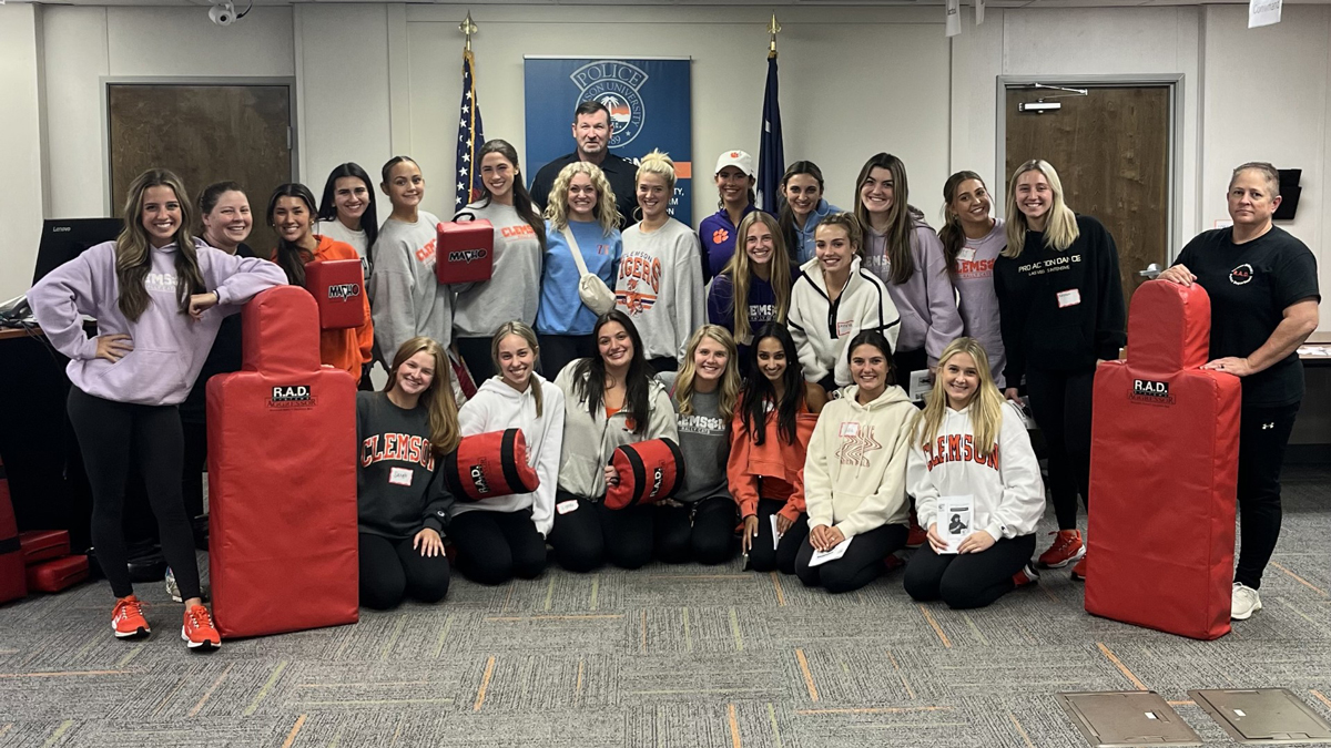 Group of students posing in a training room with red self-defense practice dummies and pads, in front of American and state flags.