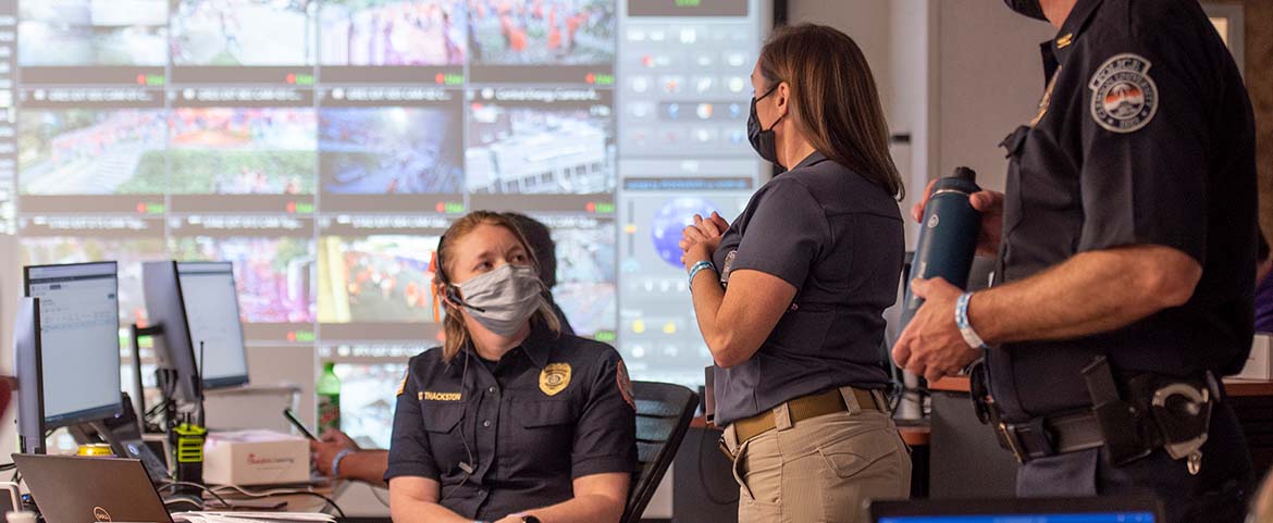 Two female emergency responders talk in front of a large screen displaying an aerial view of a crowded Clemson campus.