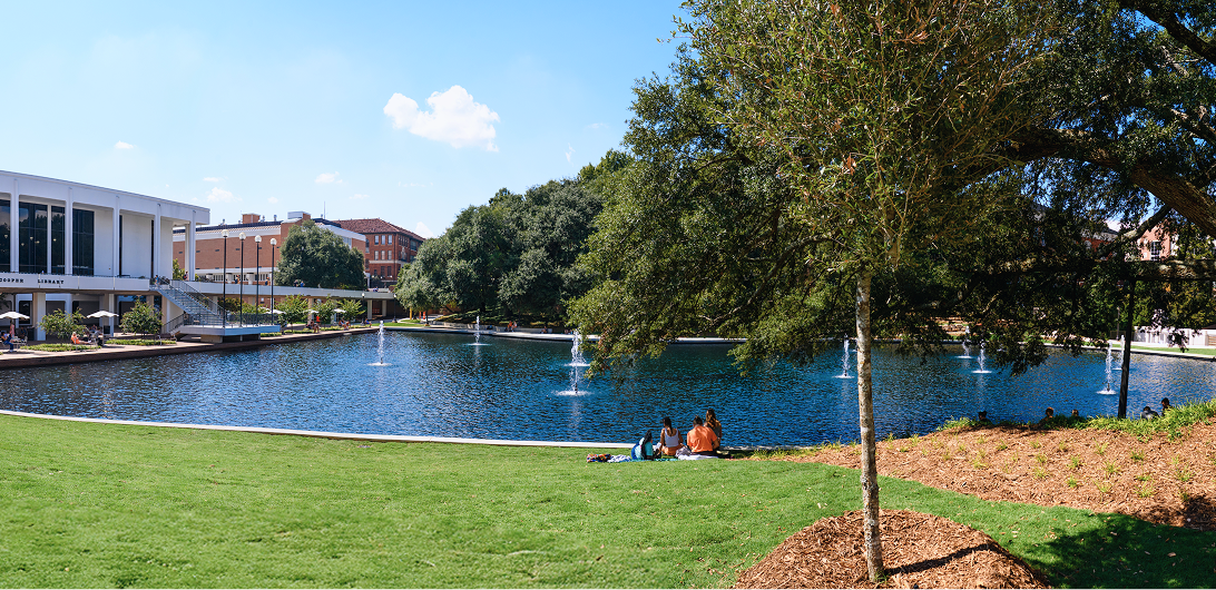 Four students lay in a grassy area in front of the Clemson University reflection pond on a sunny day.