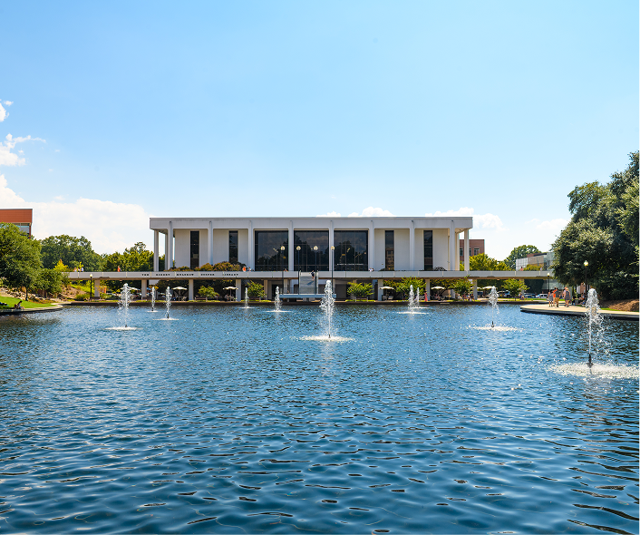 A large white white building with tall black windows sits behind a bright blue reflection pond that has several fountains spraying water above the surface of the water.