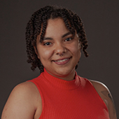 Head-and-shoulders studio portrait of a smiling person with curly hair wearing a red sleeveless top against a dark background.