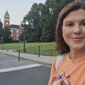 A person smiling in a casual orange t-shirt, with a university clock tower in the background.