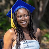 Smiling graduate wearing a blue cap with a gold tassel and a strapless white dress, outdoors with a blurred green background.