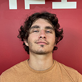 A young man with curly hair and a confident expression stands in front of a red background.