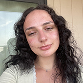 Close-up portrait of a person with long curly dark hair, freckles, and natural makeup, wearing a light top, with a blurred window and wall in the background.