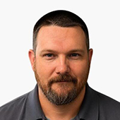 Head-and-shoulders studio portrait of a man with short dark hair and a trimmed beard, wearing a dark collared shirt against a white background.