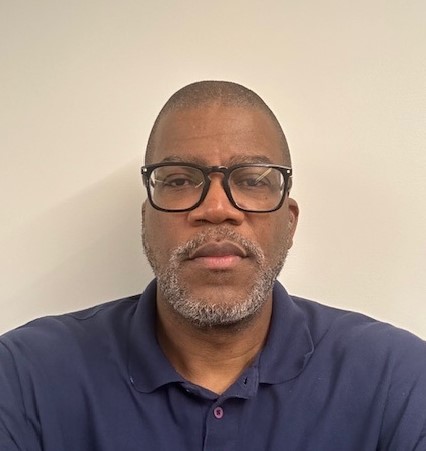 A headshot of a man wearing glasses and a navy polo shirt, with a neutral background.