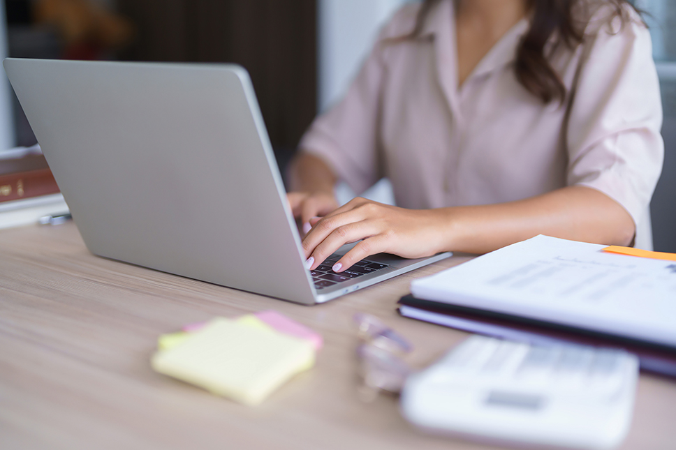 Person typing on a laptop at a desk with papers and sticky notes nearby.