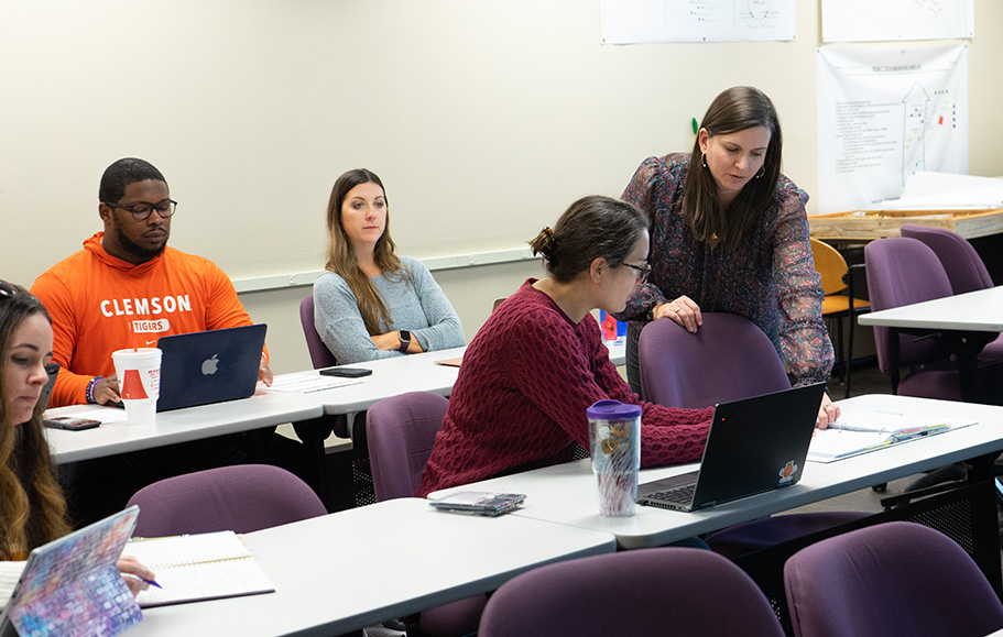 Students work in a classroom while an instructor leans over to help one student at a laptop.