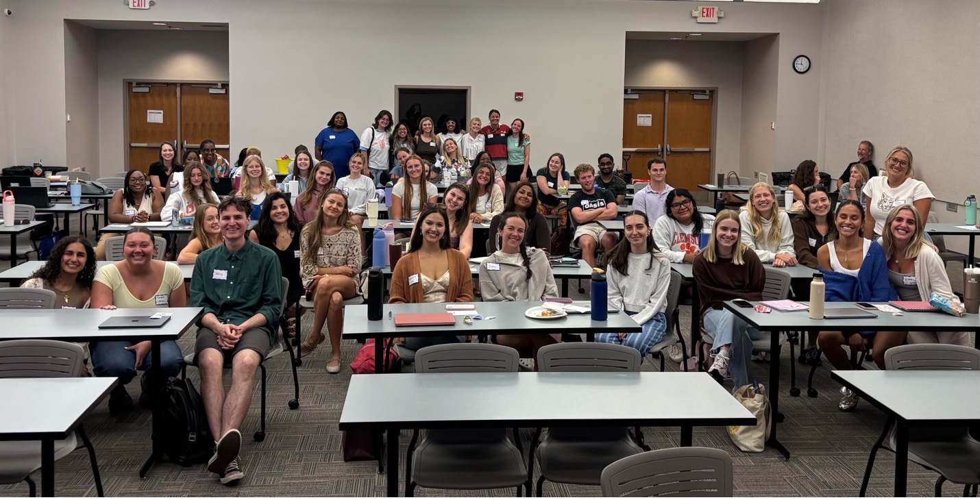 Large group of students seated at classroom tables, smiling toward the camera during a group meeting or orientation.