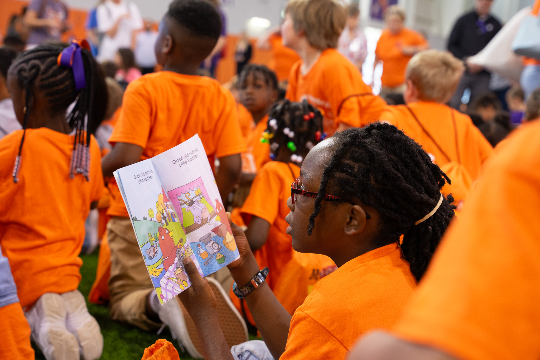 A child in an orange shirt reads a colorful book while sitting among a group of children.
