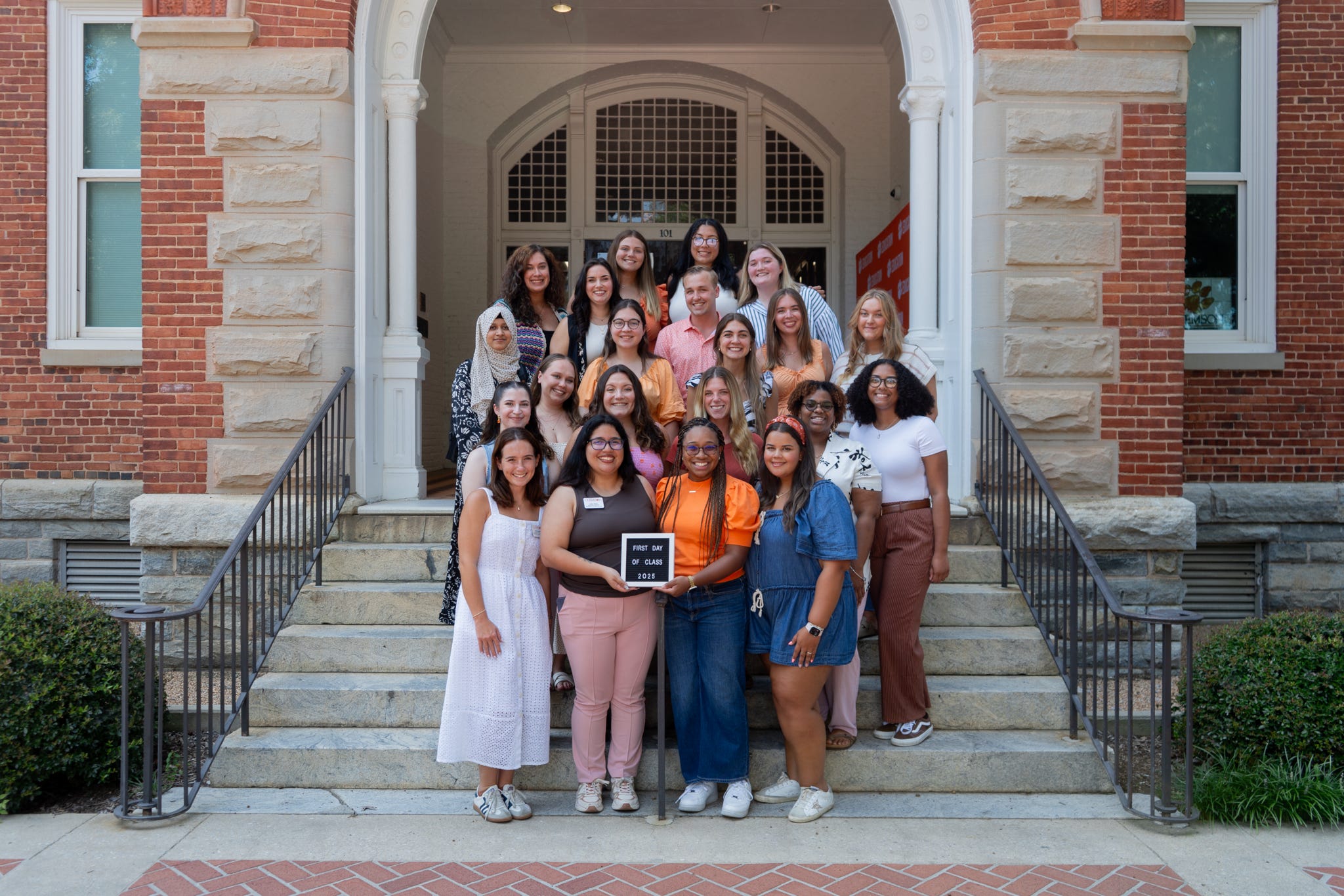 Group of students posing on the steps outside a campus building, with two holding a “First Day of Class 2025” sign.