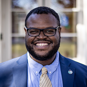 Smiling man in a suit and tie with greenery in the background.