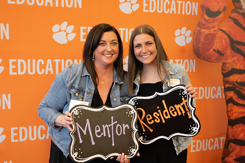 Two women pose together holding signs labeled "Mentor" and "Resident" in front of an orange backdrop with paw print and "EDUCATION" text.