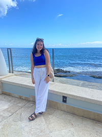 A woman in a blue blouse and white shirt stands in front of a scenic ocean background with a blue sky.
