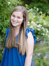 A smiling young woman with long hair, wearing a blue dress, stands in front of a lush green backdrop with flowers.