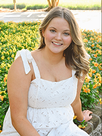 A young woman in a white dress poses outdoors with a backdrop of vibrant yellow flowers.