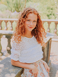 A young girl with curly red hair smiles while sitting outdoors in a floral outfit.