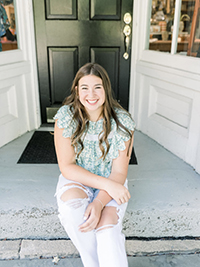 A young woman with long hair is sitting on the steps of a front door, wearing a light blue, ruffled top and ripped white jeans, smiling pleasantly.