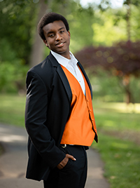 A young man in a black suit and an orange vest poses confidently outdoors.