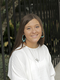 A smiling young woman with long hair, wearing a white shirt and turquoise accessories, poses outdoors near a black fence.